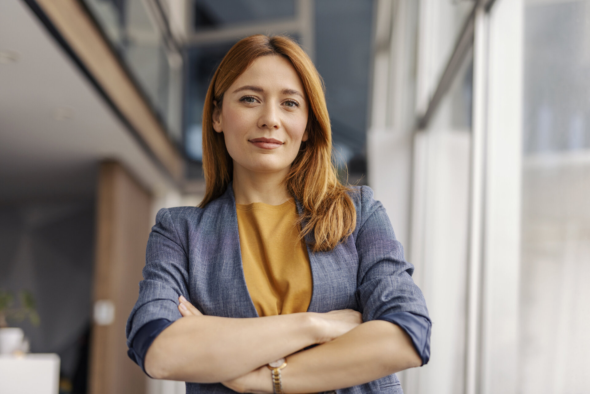 Eine Frau in blauem Blazer und gelbem Oberteil steht selbstbewusst in einem modernen Büro, mit einer Glaswand und einer weißen Wand im Hintergrund.
