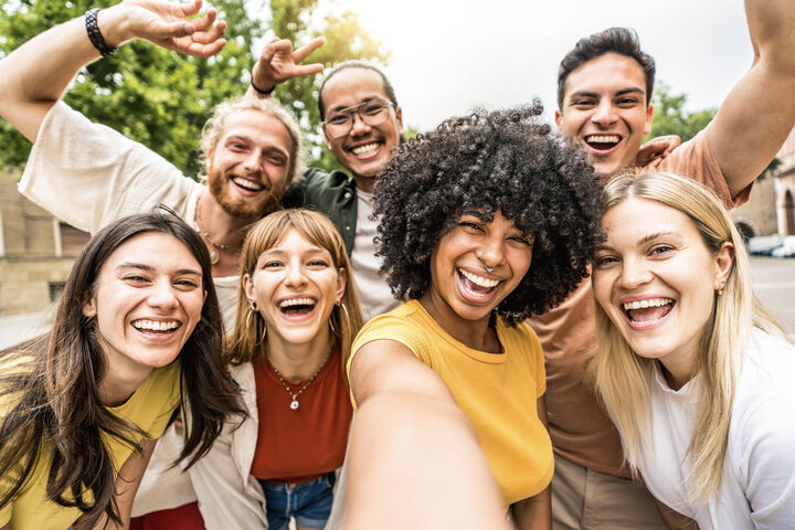 Eine Gruppe von Freunden, darunter eine Frau mit lockigem Haar und ein Mann mit Brille, lächeln und posieren für ein Selfie in einem Park.